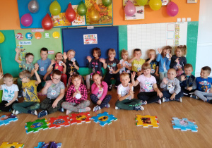 Cała grupa dzieci pozuje do wspólnego zdjęcia. Na szyi przedszkolaki maja zawieszone emblematy z napisem: Dzień Przedszkolaka. Nad dziećmi wiszą kolorowe, ozdobione balony.